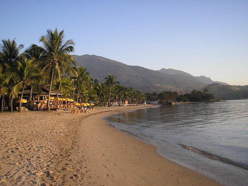 Uma praia de areia com palmeiras, guarda-sóis amarelos e uma cadeia de montanhas ao fundo sob um céu limpo.