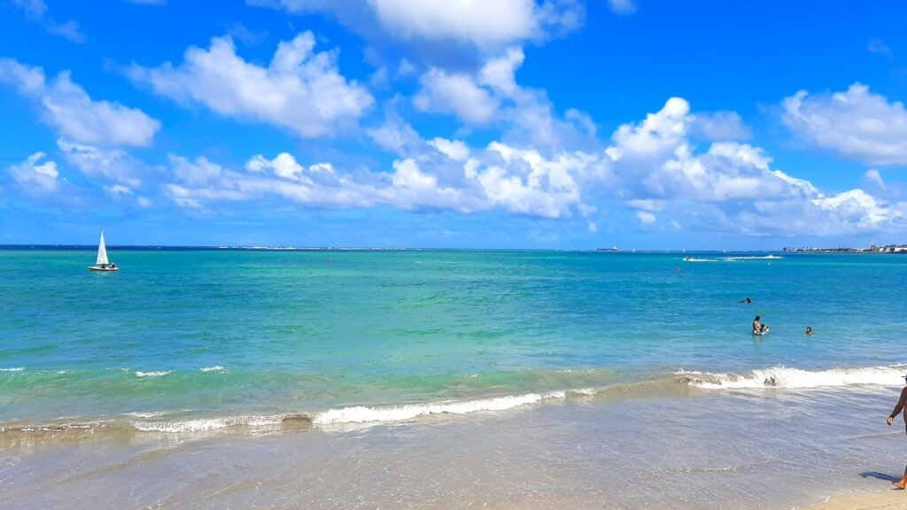 Praia em Maceió com poucas ondas, durante um dia ensolarado com poucas núvens no céu. O mar é cristalino e azul