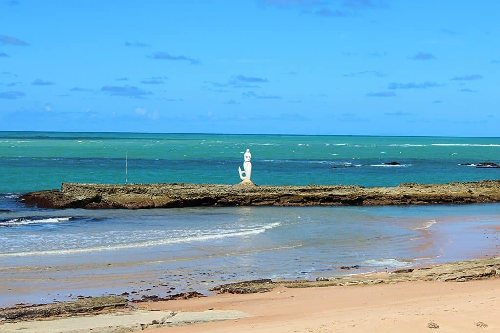 Praia da Sereia, uma das opções de o que fazer em Maceió, com águas com poucas ondas e tom azulado. No meio há uma barreira natural com uma escultura de sereia acima