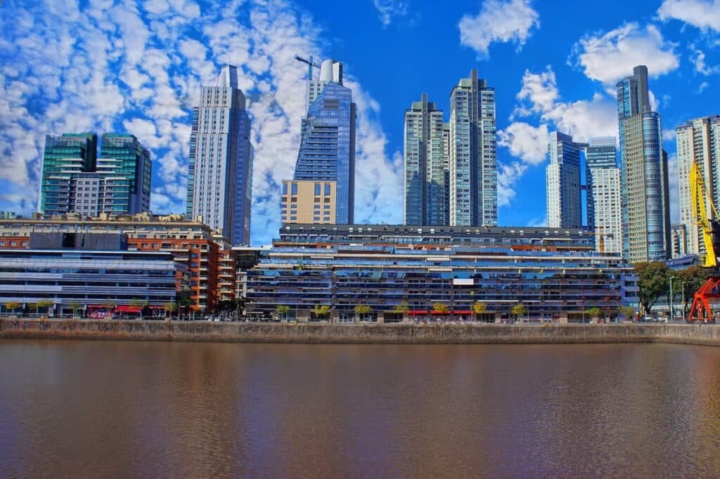 A imagem mostra a impressionante paisagem urbana de Puerto Madero, um dos bairros mais modernos e sofisticados de Buenos Aires. No primeiro plano, pode-se ver o canal que corta a área, refletindo as estruturas imponentes dos edifícios ao fundo. Os prédios são altos e modernos, com fachadas de vidro e arquitetura contemporânea, representando o desenvolvimento e a modernidade da região. O céu azul repleto de nuvens brancas complementa a beleza do local, destacando ainda mais o contraste entre a arquitetura de vanguarda e a serenidade das águas do canal. Puerto Madero é conhecido por suas construções luxuosas, e essa imagem capta perfeitamente o dinamismo e a elegância desse bairro portenho.
