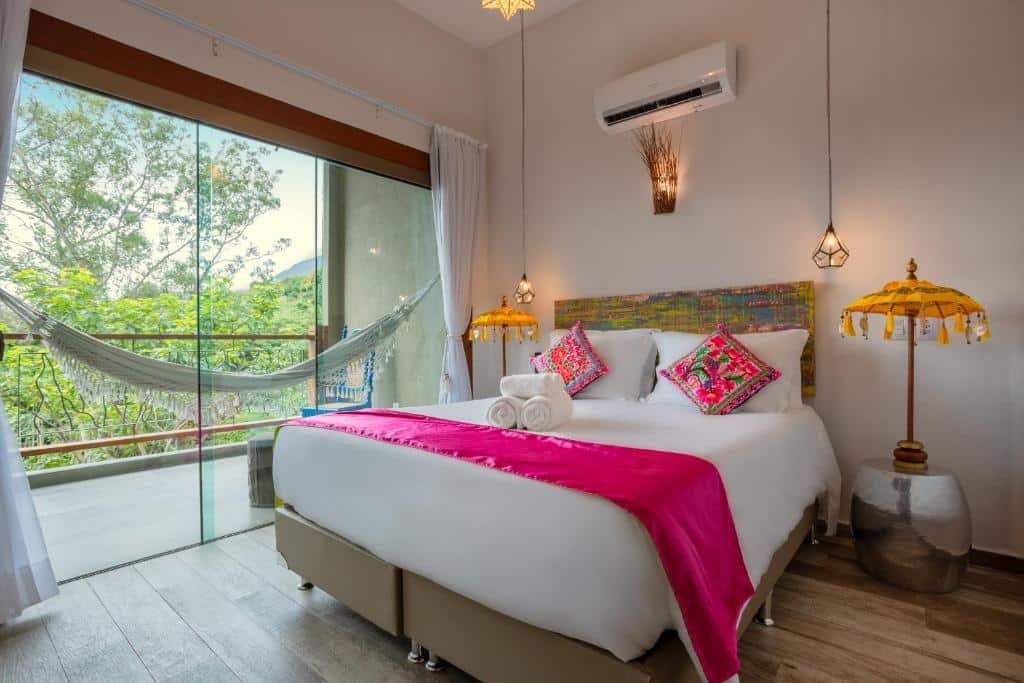 Foto do quarto em Hotel Vila Kebaya. Há uma cama box de casal no centro. Na parede atrás dela há um ar-condicionado e duas luminárias penduradas no teto. Dos dois lados da cama há um abajur. No lado esquerdo há uma porta de vidro com passagem para a varanda. Há uma rede na varanda.