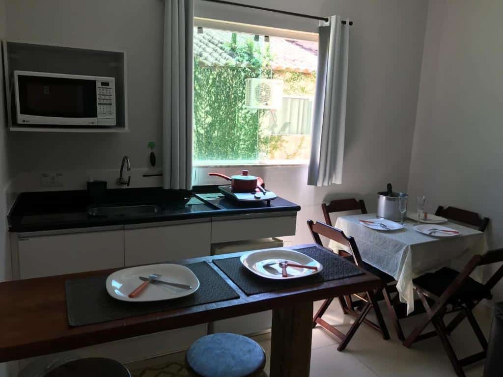 Cozinha do Residencial Camburyá. Uma bancada com pratos e talheres está em frente à pia, que tem um frigobar ao lado e um micro-ondas na parede acima. Uma janela com cortinas está logo ao lado do micro-ondas. Embaixo da janela fica uma mesinha quadrada com cadeiras.