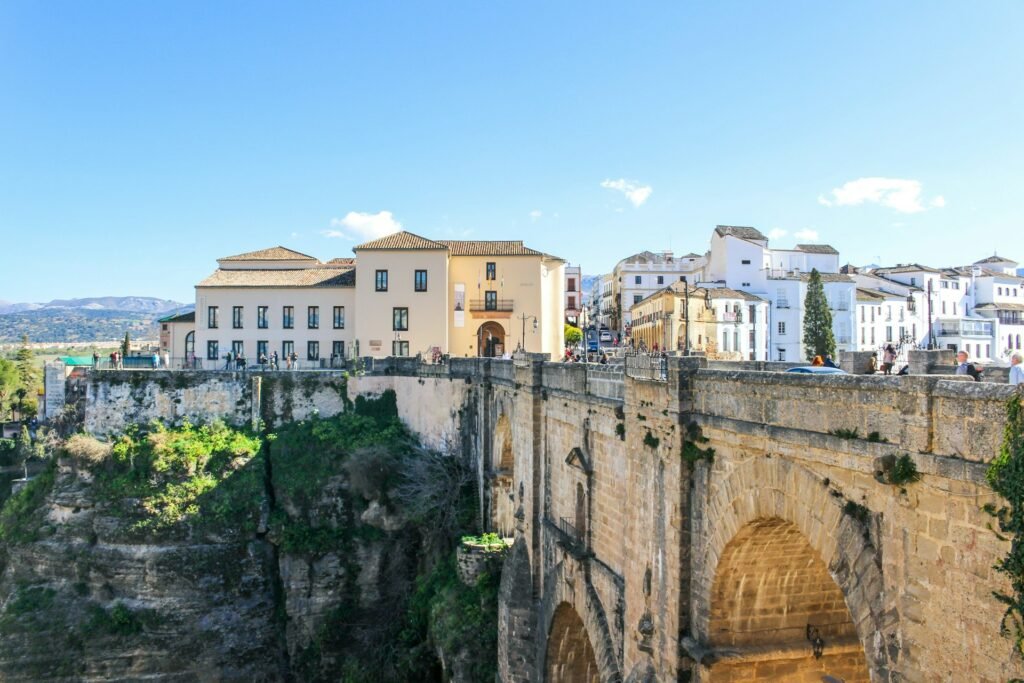 Vista de uma ponte de pedra histórica atravessando um desfiladeiro profundo em uma paisagem ensolarada, com edifícios e colinas ao fundo.