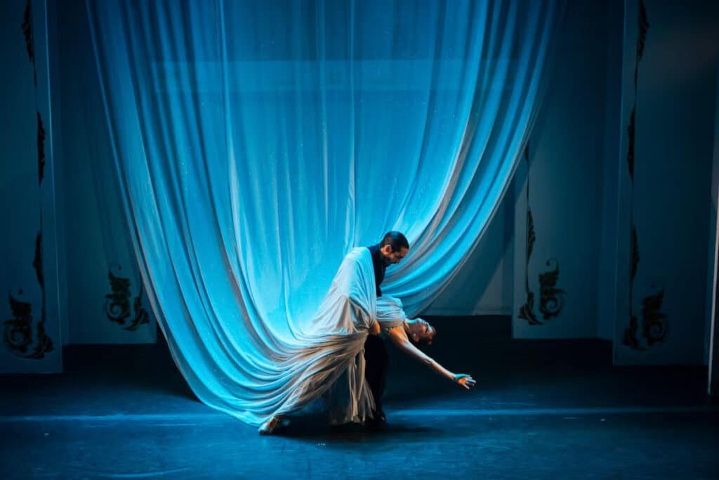 A imagem mostra um casal de dançarinos de tango em uma pose dramática e elegante em um palco, provavelmente no Café de los Angelitos, um famoso local de tango em Buenos Aires. A cena é iluminada por uma luz azul suave, que realça a fluidez e a intensidade da dança. Ao fundo, uma grande cortina drapeada envolve os dançarinos, criando a ilusão de que o vestido da dançarina se mescla ao tecido, como se fosse uma extensão do traje que desce do teto. Essa fusão entre a cortina e o vestido dá a impressão de que o vestido é ainda maior, aumentando a dramaticidade e a magia da apresentação. O efeito visual criado pela combinação do cenário com o movimento dos dançarinos torna a cena ainda mais envolvente e poética.