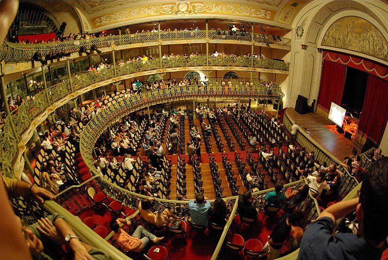 Foto tirada do interior do Teatro José de Alencar, em Fortaleza. Temos a visão de um ângulo superior, e o palco está na direita, lá embaixo. Há pessoas sentadas em assentos aleatórios, e não são todos deles que estão ocupados. O lugar é elegante, com piso de madeira e poltronas e cortinas vermelhas. Os detalhes da arquitetura são dourados e beges. É um dos pontos turísticos em Fortaleza.