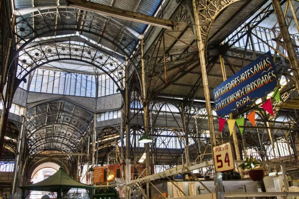 A imagem mostra o interior do Mercado de San Telmo em Buenos Aires. O teto é alto e arqueado, feito de metal e vidro, permitindo que a luz natural ilumine o espaço. A estrutura metálica do teto exibe detalhes ornamentados que remetem ao estilo arquitetônico do final do século XIX. Nas laterais, há placas indicando os tipos de produtos oferecidos, como antiguidades, artesanatos, colecionismo, curiosidades e roupas. Bandeirolas coloridas acrescentam um toque festivo ao ambiente. O mercado é vibrante e histórico, refletindo a rica cultura e diversidade de Buenos Aires.