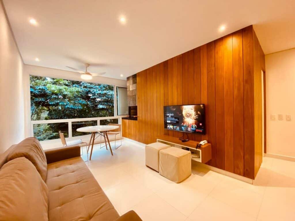 Sala de estar da Vista Mágica na Praia de Camburizinho A, onde um sofá encara uma TV com pufes e um móvel abaixo na parede oposta. Ao fundo do lado esquerdo há uma mesa em frente à churrasqueira e com uma longa janela com vista ao lado.