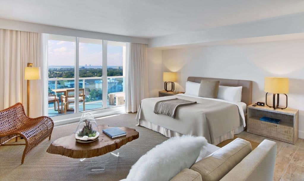 A imagem mostra um quarto decorado em tons neutros no 1 Hotel Miami. Há uma cama de casal grande, coberta com roupa de cama bege e travesseiros brancos. Duas mesas de cabeceira com lâmpadas modernas estão ao lado da cama. À esquerda, há uma poltrona de vime e uma luminária de pé. No centro do quarto, uma mesa de centro rústica de madeira exibe um terrário e um livro. Grandes janelas de vidro oferecem uma vista panorâmica da cidade e da varanda, onde há uma mesa com cadeiras.