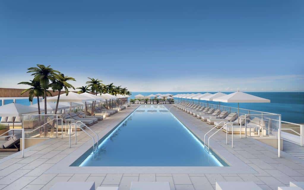 A imagem mostra uma piscina infinita em um terraço com vista para o mar no 1 Hotel Miami, um dos hotéis em Miami Beach indicados. A piscina é longa e retangular, com bordas iluminadas por luzes subaquáticas. Em ambos os lados da piscina, há fileiras de espreguiçadeiras brancas, cada uma coberta por grandes guarda-sóis brancos. Algumas palmeiras estão dispostas ao longo das bordas, adicionando um toque tropical ao ambiente. O céu está claro e azul, criando uma sensação de tranquilidade e luxo.