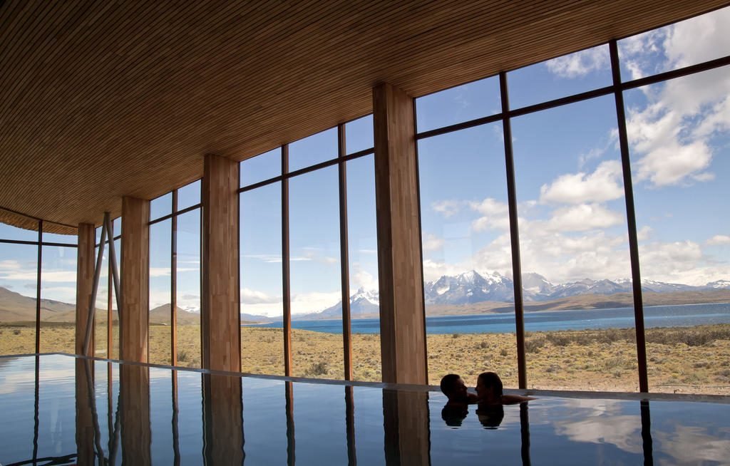 Piscina interna com grandes janelas com vista para uma paisagem cênica de montanhas, um lago e um céu parcialmente nublado, com duas pessoas dentro.