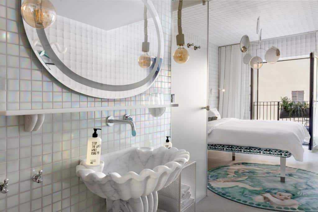 Quarto plus médio do 25hours Hotel, de 21 m², mostrando a pia do banheiro de mármore no formato de uma colcha, com um espelho redondo em cima, parede de azulejos pequenos, um produto para lavar as mãos na pia, além da cama do quarto, com um enfeite cheio de espelhos redondos suspenso no teto e uma varanda com flores suspensas na grade