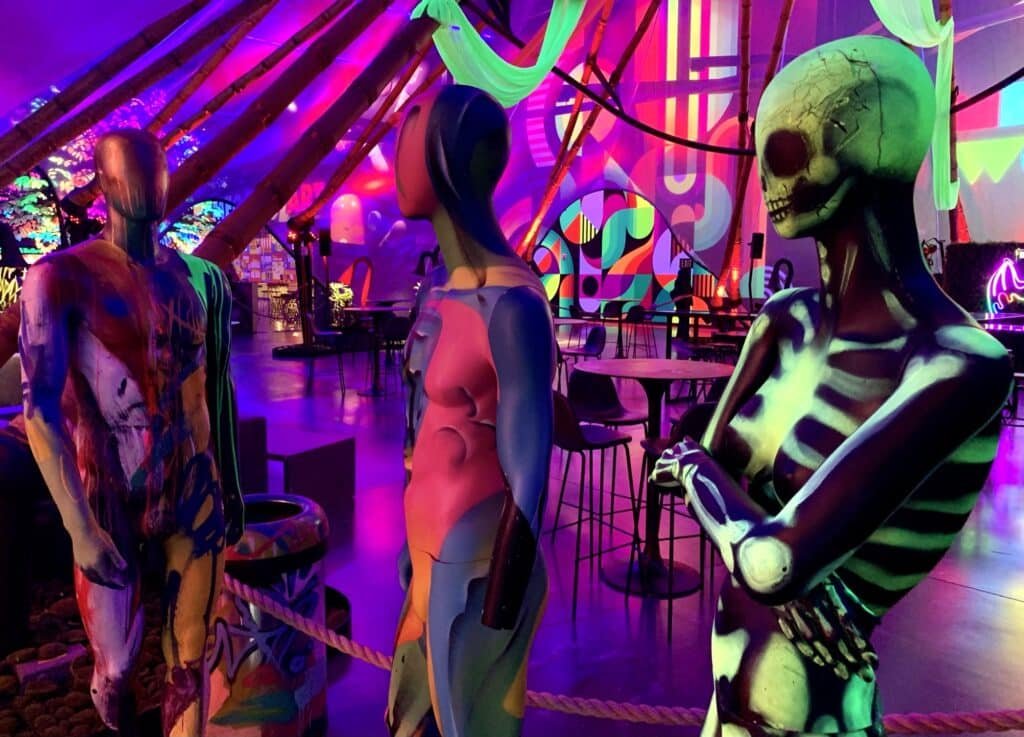 A foto mostra bonecos manequins pintados e, ao fundo, um ambiente com várias cores diferentes, panos e decorações neon. 