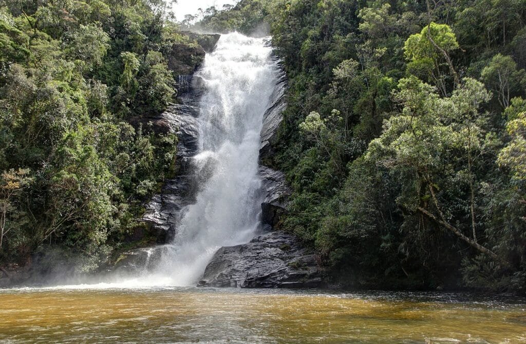 Cachoeira caindo em cascata por uma encosta rochosa cercada por vegetação densa formando uma piscina de água. Essa é a Cachoeira Santo Isidro, acessada por uma das trilhas em SP.