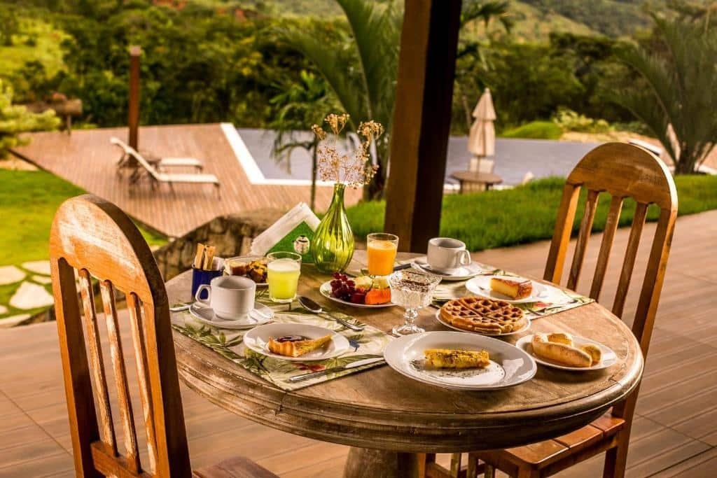 Mesa com café da manhã do hotel Solar da Serra Tiradentes. A mesa e as cadeiras são de madeira, e sobre ela há pães, waffles, frutas, entre outros. No fundo da foto é possível ver o deck da piscina e algumas espreguiçadeiras.