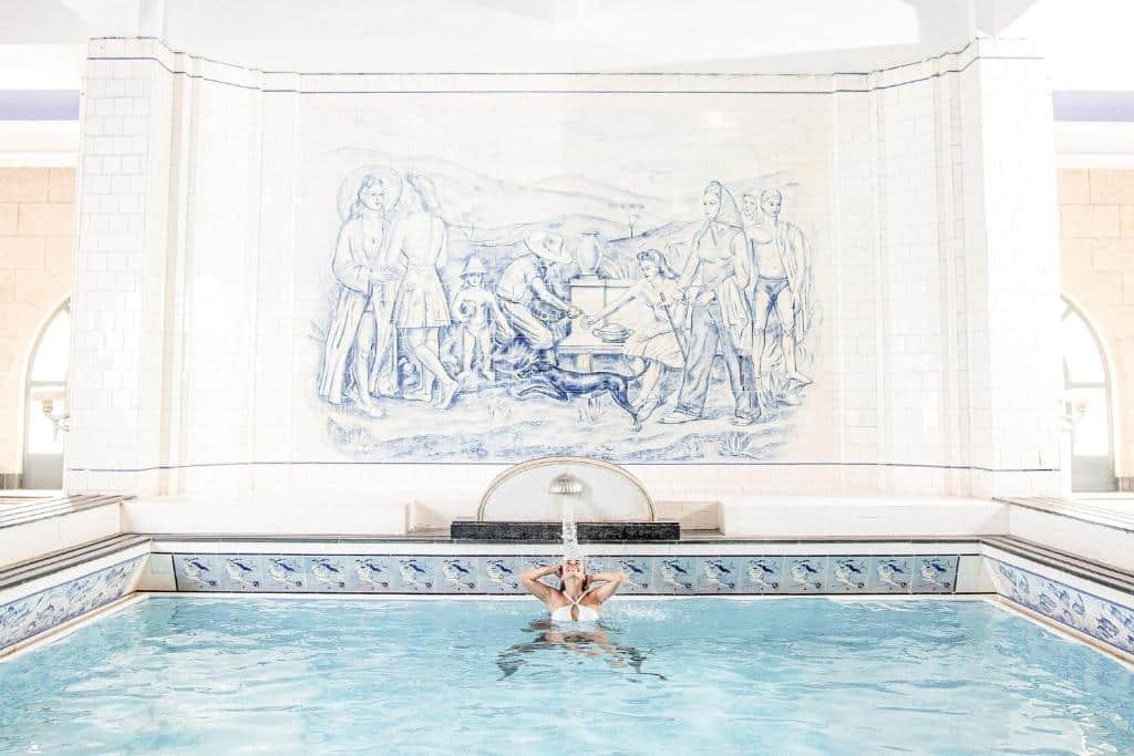 Imagem da piscina do Grande Hotel Termas de Araxá. Há um grande mural com uma pintura clássica/renascentista, os azulejos da piscina são decorados e ela possui um pequeno chafariz, onde há uma mulher molhando seus cabelos embaixo.