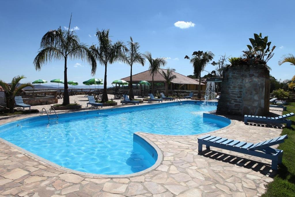 Foto da piscina do Hotel Alpha Ville Chalés. Ela é grande e ocupa um largo espaço do ambiente, e possui uma cascata de água. Vemos várias espreguiçadeiras, mesas com guarda-sol, árvores e um quiosque no fundo.