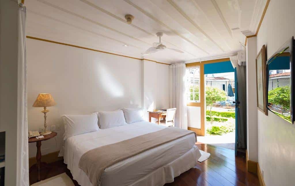 Quarto do Hotel Solar do Rosário. A cama é de casal e possui lençóis e travesseiros brancos. Do lado esquerdo, vemos uma mesa de cabeceira com um abajur e telefone em cima. Do outro lado, está uma escrivaninha e a janela do quarto, que também é uma porta com passagem para o jardim. Foto para representar o post de hotéis com piscina aquecida em Minas Gerais.