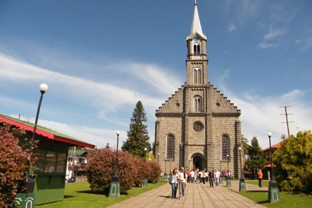 A imagem mostra a Igreja Matriz de São Pedro, localizada em Gramado, Rio Grande do Sul. A igreja é feita de pedra, com uma arquitetura em estilo gótico, caracterizada por suas linhas verticais e a torre central alta com um telhado pontiagudo. À frente da igreja, há um caminho de pedras ladeado por arbustos floridos e postes de iluminação, que leva até a entrada principal. Várias pessoas estão caminhando ou tirando fotos em frente à igreja, o que sugere que é um local turístico popular. O céu está claro e azul, contribuindo para a beleza cênica da cena.