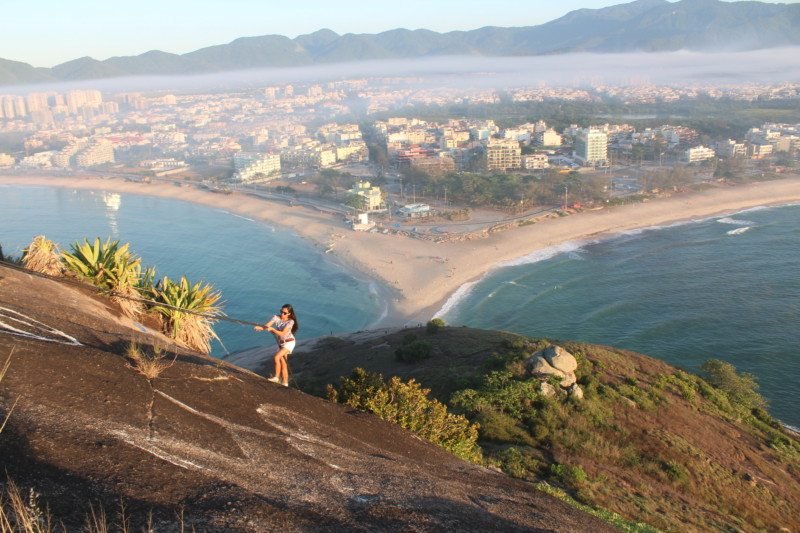 Mulher em pé em uma colina rochosa com vista para uma cidade costeira, com uma praia, oceano e montanhas visíveis ao fundo. Essa é a paisagem de uma das trilhas no Rio de Janeiro.