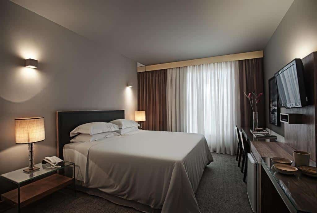 Quarto do Ouro Minas Hotel. O ambiente é moderno, a cama é de casal e seus lençóis e travesseiros são brancos. Em ambos lados, há uma mesa de cabeceira de vidro com um abajur em cima. Na frente, há a televisão suspensa na parede, um frigobar e uma mesa com cadeiras. A janela está do lado da cama. Foto para ilustrar o post sobre hotéis com piscina aquecida em Minas Gerais.