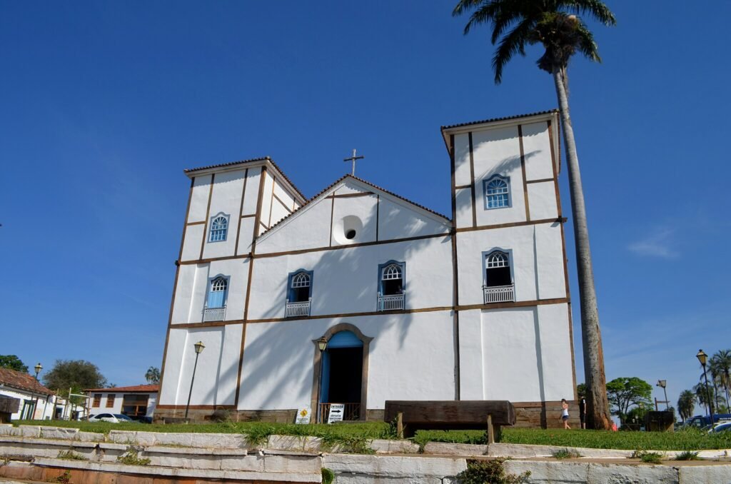 A imagem mostra a Igreja Matriz de Nossa Senhora do Rosário, localizada em Pirenópolis, Goiás, Brasil. A igreja é um edifício colonial com paredes brancas e detalhes em madeira marrom, apresentando um estilo arquitetônico tradicional. A fachada possui três grandes portas e janelas com molduras azuis, além de duas torres laterais que se destacam pela sua simetria. Em frente à igreja, há uma área gramada com um banco de madeira e alguns postes de iluminação. Ao lado direito da imagem, uma alta palmeira complementa a paisagem. O céu está limpo e azul, proporcionando um belo contraste com a estrutura branca da igreja. Algumas pessoas podem ser vistas ao redor.