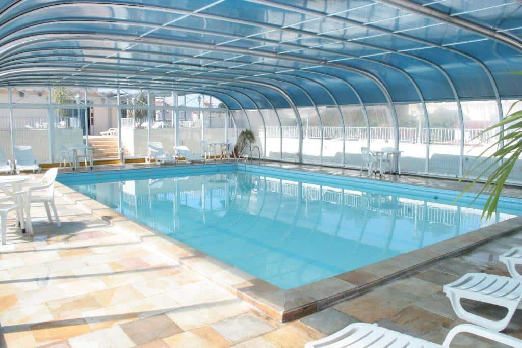 Piscina coberta do Hotel Negreiros. A área é bem iluminada, e há algumas mesas com cadeiras e espreguiçadeiras no ambiente.
