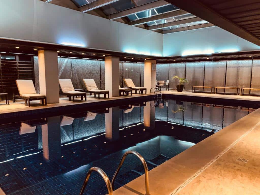 Área da piscina no Ouro Minas Hotel. A piscina é grande, há algumas espreguiçadeiras e bancos. O ambiente é bem iluminado, com luzes em cima das espreguiçadeiras e o teto possui claraboias.