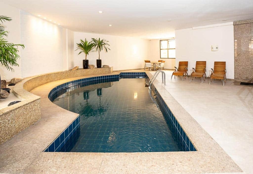 Piscina interior do Mercure Belo Horizonte. Há algumas espreguiçadeiras de madeira e uma mesa no fundo com algumas cadeiras podem ser vistas. Vasos com plantas decoram o ambiente.