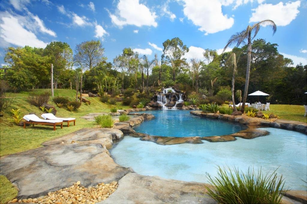 Piscina termal do Ville Real Hotel. Há muitas áreas verdes em volta, e uma pequena cachoeira que deságua em uma das piscinas. Podemos ver espreguiçadeiras e mesas com guarda-sóis no fundo.