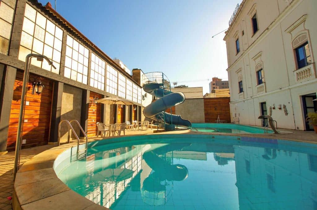 Piscina do Palace Hotel de Caxambu. Há duas piscinas diferentes, uma é mais rasa e possui um escorregador aquático. Algumas mesas com cadeiras e guarda-sol estão disponíveis para uso.