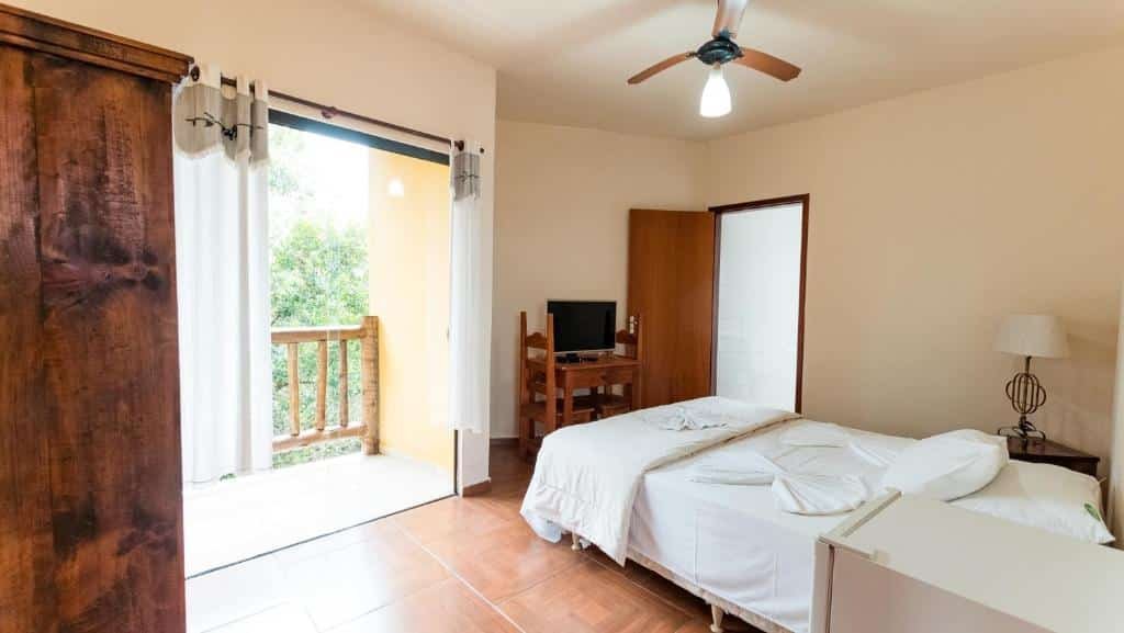 Quarto do Hotel Alpha Ville Chalés. A cama é box e de casal, e está encostada na parede com vista para a janela. Há uma porta de vidro que dá acesso à varanda da suíte. De frente para a cama, há um suporte de madeira para a TV, e do lado está a porta. Foto para ilustrar o post sobre hotéis com piscina aquecida em Minas Gerais.