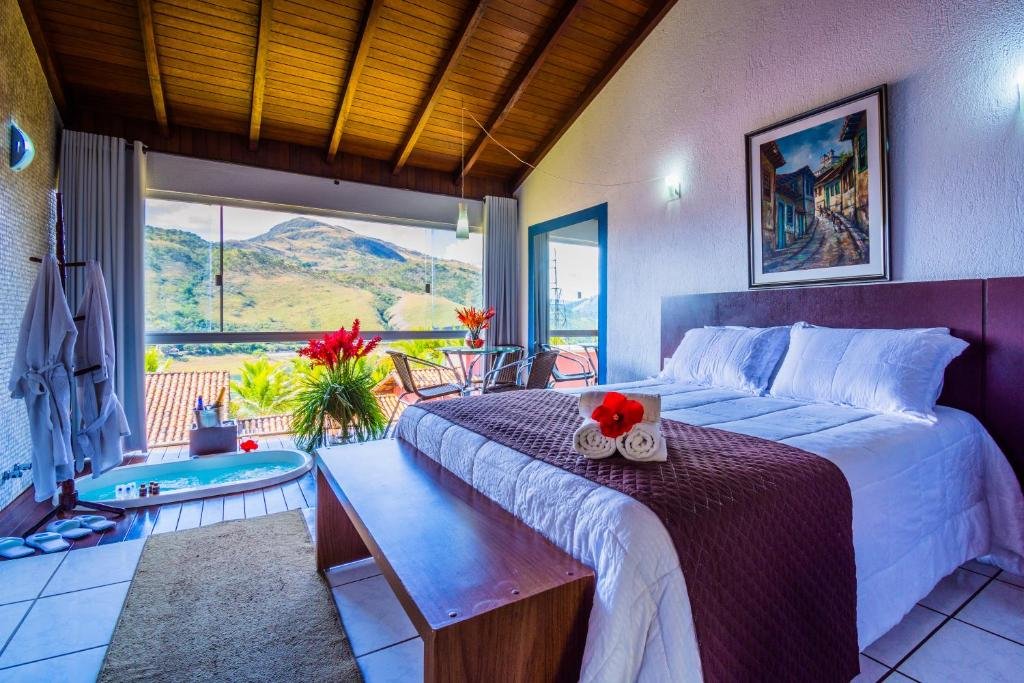Quarto do Balneário do Lago Hotel. Foto para representar o post sobre hotéis com piscina aquecida em Minas Gerais. A cama é box e de casal, na sua frente há um banco de madeira. Do lado, há uma mesa com cadeiras, plantas e uma banheira de hidromassagem. Há uma varanda no quarto e um cabide com roupões pendurados.