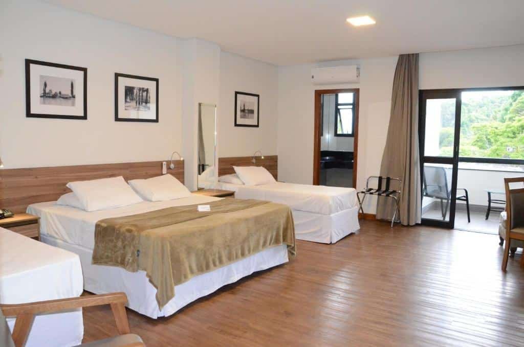 Quarto do Hotel Metrópole. A suíte é espaçosa e há uma cama de casal entre outras duas camas de solteiro. É possível ver três quadros em preto e branco decorando a parede. Do lado de uma das camas, há uma porta para o banheiro do quarto. Há outra porta que dá passagem para a varanda. Foto para ilustrar o post sobre hotéis com piscina aquecida em Minas Gerais.