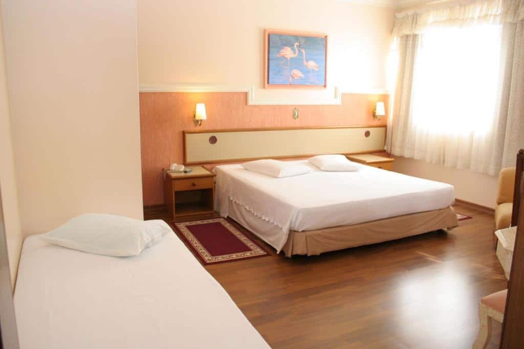 Quarto do Hotel Negreiros. A foto ilustra o post sobre hotéis com piscina aquecida em Minas Gerais. Há uma cama box de casal e outra de solteiro na suíte. Na de casal, há uma mesa de cabeceira em cada lado, e abajures fixos na parede. Um quadro decora um pouco à cima. Uma janela está ado lado direito.