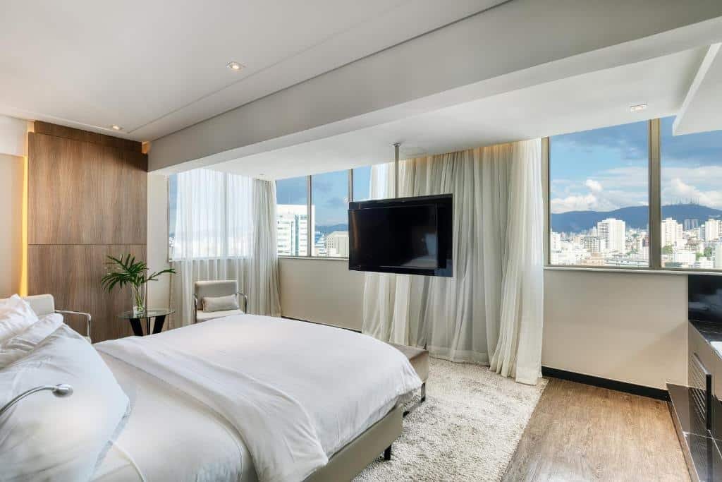 Foto do Mercure Belo Horizonte. A imagem foi tirada em direção da cama para o restante do quarto, sendo possível ver as janelas que dão vista para a cidade, e a televisão, que está suspensa por uma barra de ferro no teto. Imagem ilustrativa do post de hotéis com piscina aquecida em Minas Gerais.