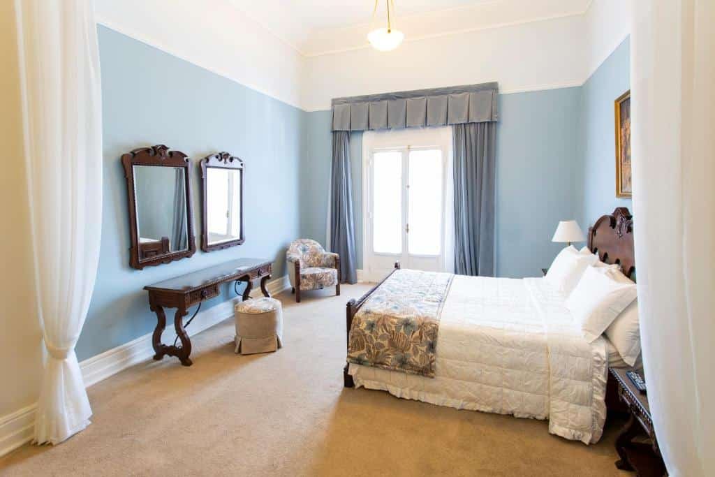 Foto do quarto do Palace Hotel - Poços de Caldas. Os móveis possuem um estilo antigo, dando um ar mais clássico para o ambiente. A cama é de casal e sua cabeceira é de madeira escura. Na parede em que a mesma está encostada há um fixo quadro decorando a suíte. De frente para a cama, há uma pequena mesa de madeira com gavetas, dois espelhos e um puff. Do lado esquerdo da cama, há uma grande janela com cortinas azuis claras, e uma poltrona ao seu lado. Foto para ilustrar o post sobre hotéis com piscina aquecida em Minas Gerais.