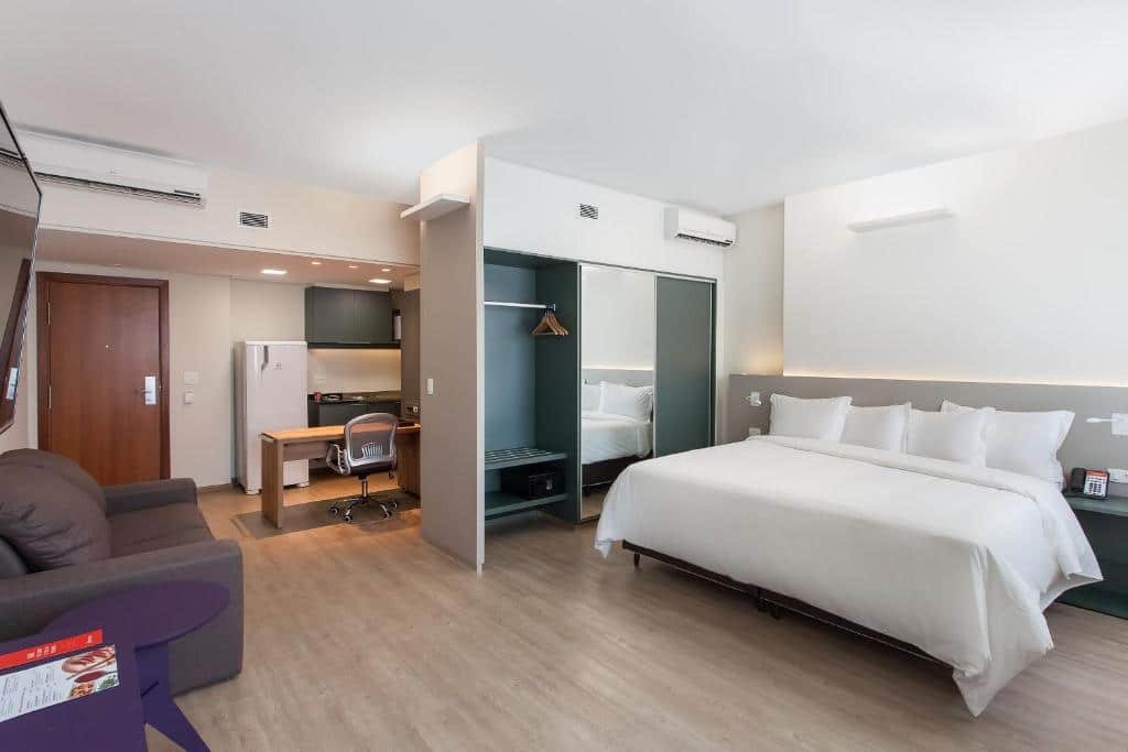 Quarto do Ramada Encore by Wyndham. A cama é box e larga, com vários travesseiros e lençóis brancos. Do lado esquerdo, há um guarda-roupa de três portas. Na frente da cama, vemos uma parte da televisão que está fixa na parede. Há um sofá na mesma parede da TV. Atrás da parede do guarda-roupa que separa duas partes do quarto, há uma pequena cozinha equipada. Imagem par representar o post sobre hotéis com piscina aquecida em Minas Gerais.