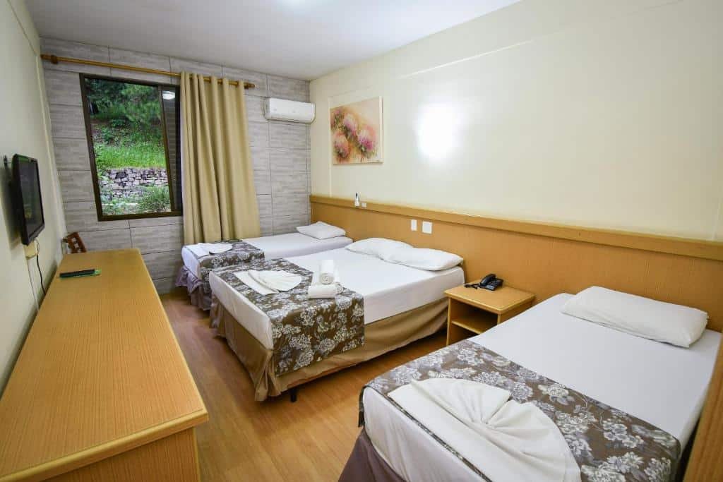 Foto do Vilage Inn All Inclusive Poços de Caldas. Há uma cama box de casal entre outras duas camas de solteiro. Na frente, há uma mesa de televisão, e a TV está fixa na parede. O quarto possui uma janela e um ar-condicionado do lado.