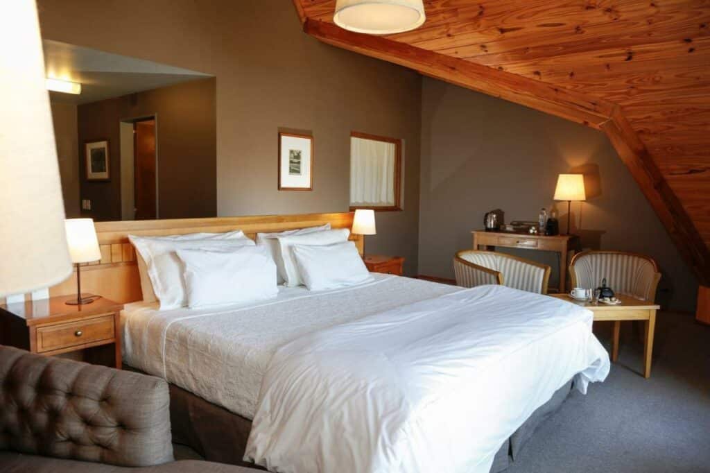 Quarto do Los Cerros del Chaltén Boutique Hotel com cama de casal do lado esquerdo da imagem com uma cômoda em cada lado com luminária e do lado esquerdo da cama duas cadeiras com mesa. Representa hotéis em El Chalten.