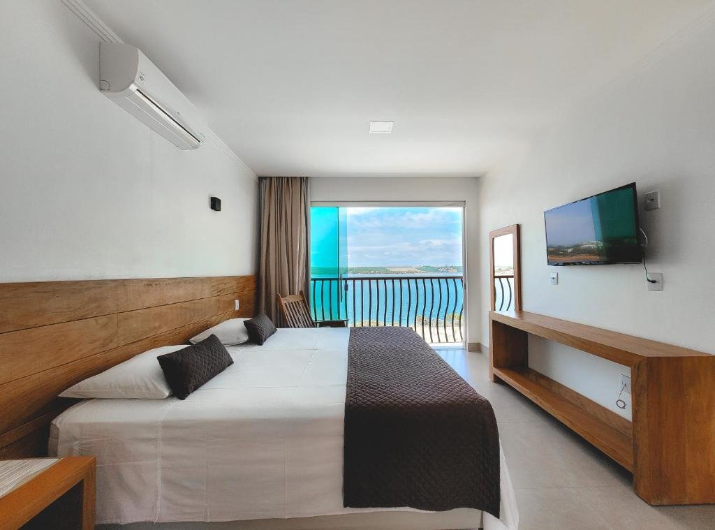 Quarto do hotel Riviera Capitólio Hotel. A cama de casal está encostada na parede esquerda. De frente, há uma mesa de madeira, e a TV está fixa na parede. A janela está no centro da imagem, e sua cortina é marrom. Imagem para representar o post de hotéis com piscina aquecida em Minas Gerais.