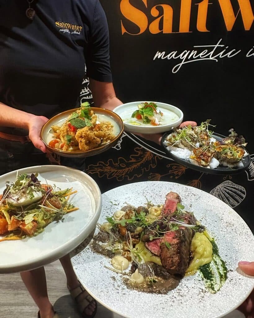 Uma pessoa segurando cinco pratos gourmet, incluindo salada, macarrão e carnes grelhadas, no Saltwater Restaurant Magnetic Island.