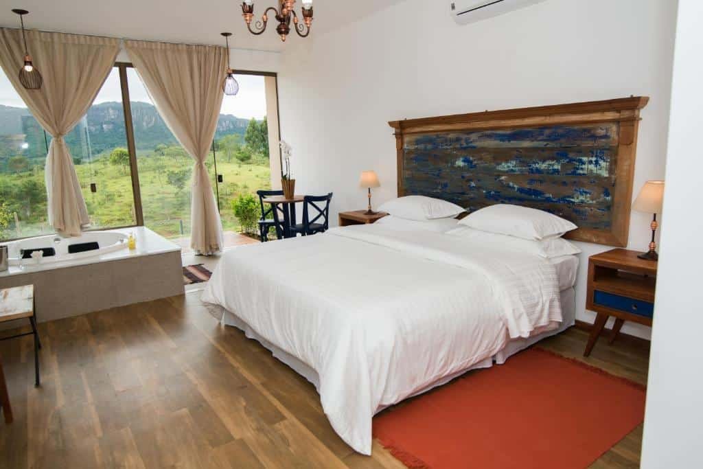Quarto do hotel Solar da Serra Tiradentes. A cama do hotel possui lençóis e travesseiros brancos, e sua cabeceira e mesas de canto são rústicas. Elas tem dois abajures um em de cada. O tapete do quarto é vermelho. Na esquerda, vemos uma pequena mesa circular com duas cadeiras azuis de madeira, e um vaso com flores em cima. A janela é grande e percorre toda extensão do quarto, as cortinas são beges. Uma hidromassagem está disponível para uso do lado da janela. Imagem para representar o post de hotéis com piscina aquecida em Minas Gerais.
