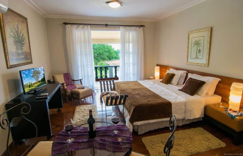 Quarto do Garden Hill Hotel e Golfe. A cama é de casal e bem espaçosa e está encostada na parede. Nos lados, há mesas de cabeceiras almofadadas com abajur em cima. De frente para a cama, há um pequeno sofá-cama, e uma mesa de televisão. De um lado do quarto, há uma mesa de vidro e há um vinho e taças em cima. Do outro, há uma poltrona e uma janela com varanda.