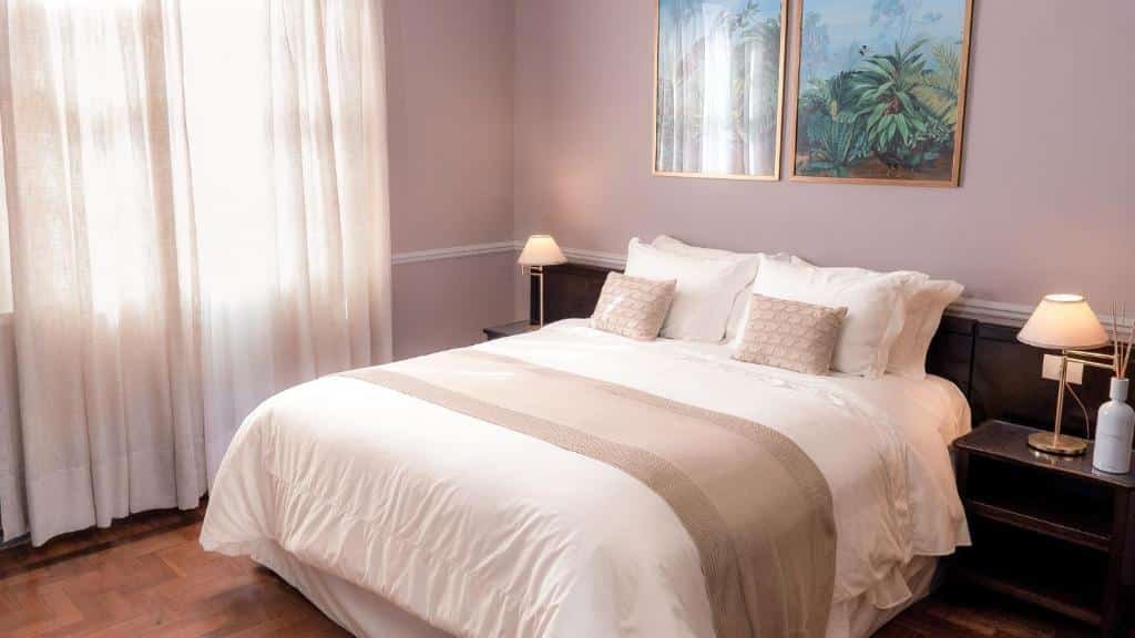 Quarto do Grande Hotel Termas de Araxá. A cama é de casal e possui lençóis e travesseiros claros. Há uma mesa de escrivaninha com um abajur de cada lado da cama, uma delas também possui um incenso em cima. Dois quadros decoram a mesma parede na qual a cama está encostada. A janela do quarto está do lado esquerdo, e possui cortinas brancas. Foto para ilustrar o post sobre hotéis com piscina aquecida em Minas Gerais,