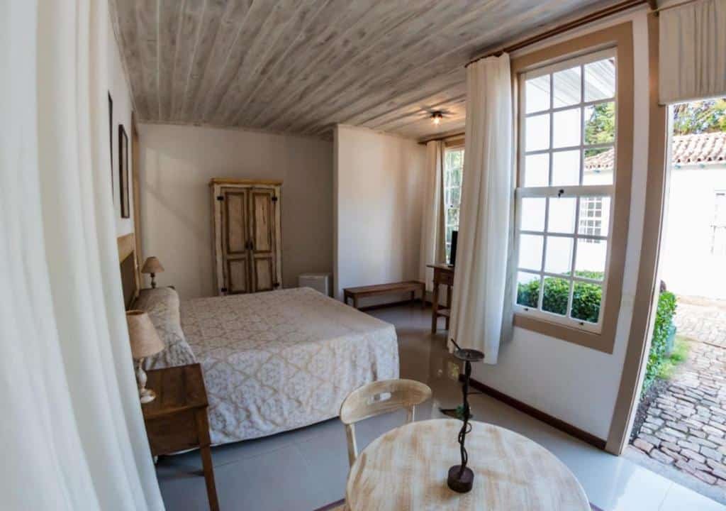 Suíte do Ville Real Hotel. A decoração do quarto é colonial. A cama de casal está no centro, e duas mesas de cabeceira de madeira com abajur estão de ambos os lados. Há um pequeno armário de madeira encostado na parede do lado da cama. Do outro lado, é possível ver parte de uma pequena mesa de madeira redonda com cadeiras. Há janelas e uma porta que dá passagem para um jardim. Foto para ilustrar o post sobre hotéis com piscina aquecida em Minas Gerais.