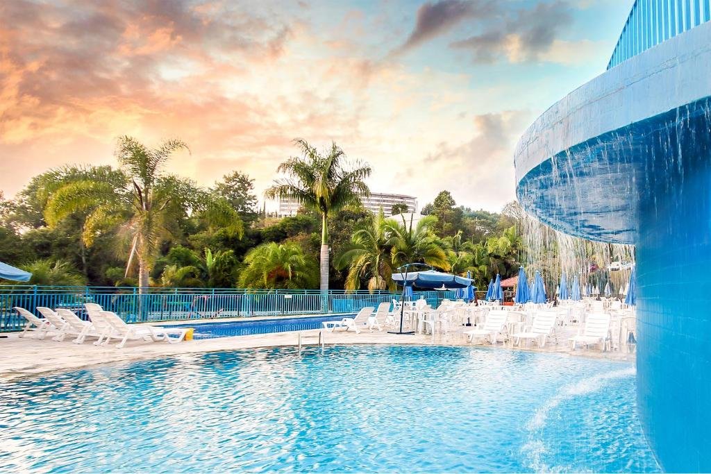 Área da piscina do Vilage Inn All Inclusive Poços de Caldas. A piscina é grande, e há mais de uma. Muitas espreguiçadeiras e mesas com guarda-sol estão no ambiente. Uma grande cascata de água está no meio de uma das piscinas.