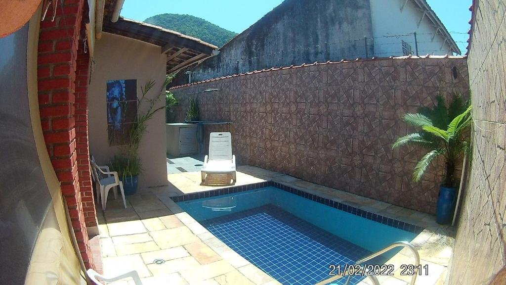 Área externa da A Casa do Chef Ubatuba. Uma piscina no lado direito, no fundo uma cadeira de praia, do lado esquerdo uma cadeira e um vaso de planta e a casa.