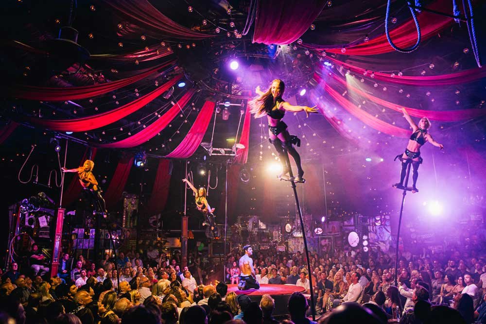 Uma apresentação de circo do Absinthe com acrobatas equilibrando-se em postes acima de um público lotado, sob uma tenda com cortinas vermelhas e iluminação brilhante.