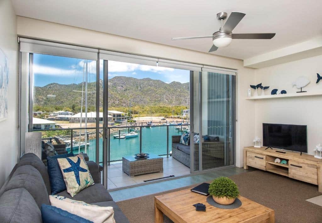 Sala de estar do Absolute Waterfront Magnetic Island com ventilador de teto, sofás cinza, mesa de centro de madeira, TV e portas de vidro com vista para a marina e as montanhas.