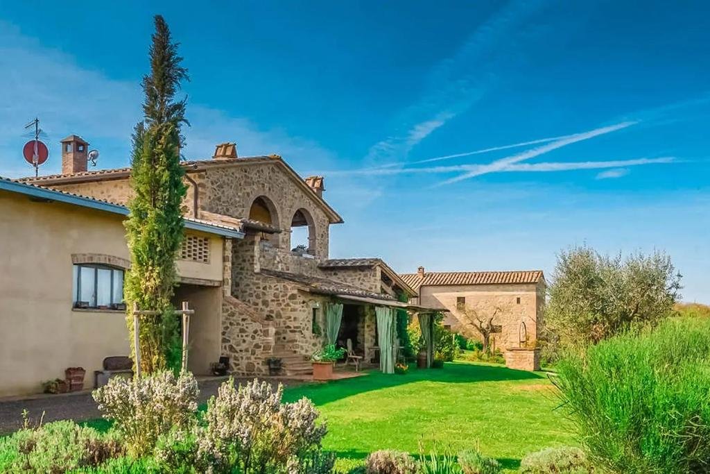 Área externa da casa de temporada Agriturismo Monsole, com gramado verde baixo, algumas árvores verdes e flores, e entrada da casa com algumas cadeiras na frente